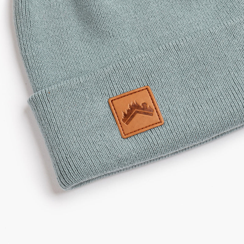 A close up detail shot of a soft glacier blue 6 panel beanie with a custom ALDRI SUR leather logo patch square stitched to the bottom left fold