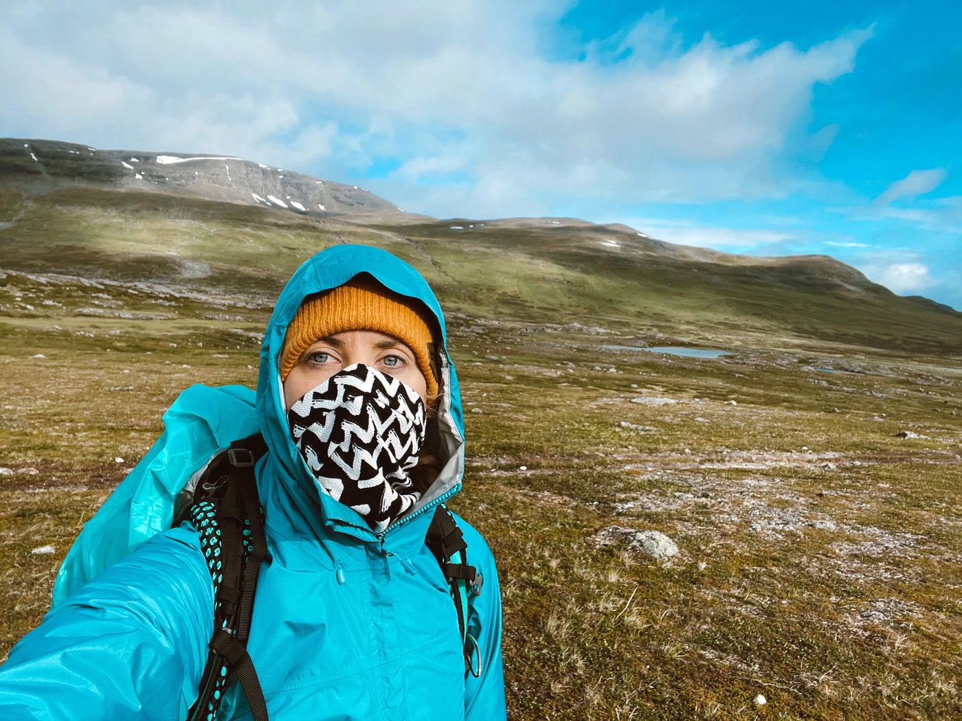 A hiker wearing a black and white zig zag pattern face covering neck gaiters standing in front of Swedish tundra in Lapland with an all turquoise raincoat and trekking pack and a rust orange knitted wool beanie made by ALDRI SUR
