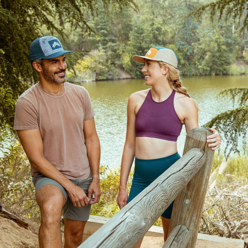 Lightly bearded man wearing Dark Blue Aldri Sur Topo Performance Running Hat, t shirt, and shorts smiling at blonde woman in a purple sports bra, tight fitness compression shorts, and a Teal Orange Tan 5 Panel Friluft Cotton Camper hat by Aldri Sur. Both are in front of a lush green forest and lake in the summer time