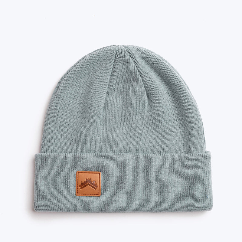 A soft glacier blue 6 panel beanie with a custom ALDRI SUR leather logo patch square stitched to the bottom left fold