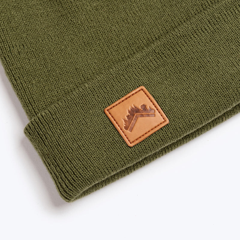 A close up detail shot of a soft recycled acrylic moss green 6 panel beanie with a custom ALDRI SUR leather logo patch square stitched to the bottom left fold