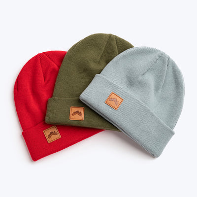A trio flat lay shot of the ALDRI SUR recycled acrylic beanie in Spice Red, recycled acrylic beanie in Moss Green, and cotton acrylic blend in soft Glacier Blue. All have custom leather logo patch squares in the bottom left corner of the fold