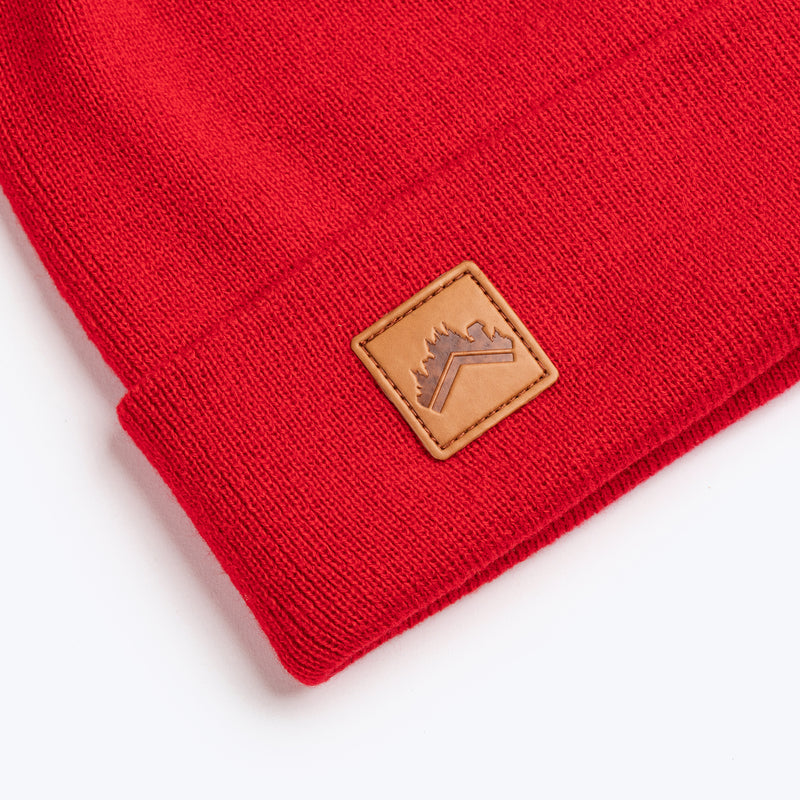 A close up detail shot of soft spice red recycled acrylic 6 panel beanie with a custom ALDRI SUR leather logo patch square stitched to the bottom left fold