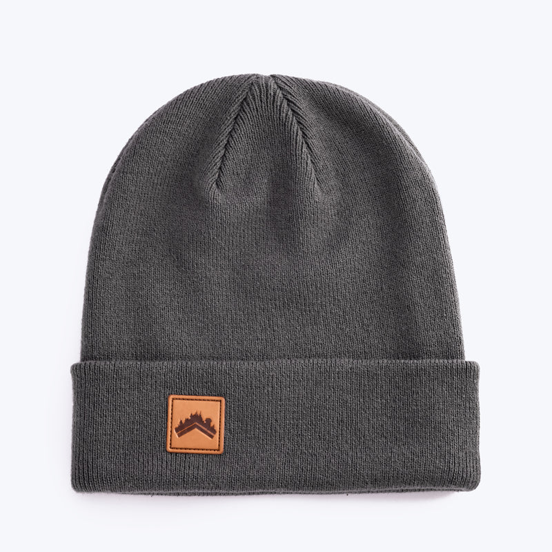 A soft dark stone grey 6 panel beanie with a custom ALDRI SUR leather logo patch square stitched to the bottom left fold