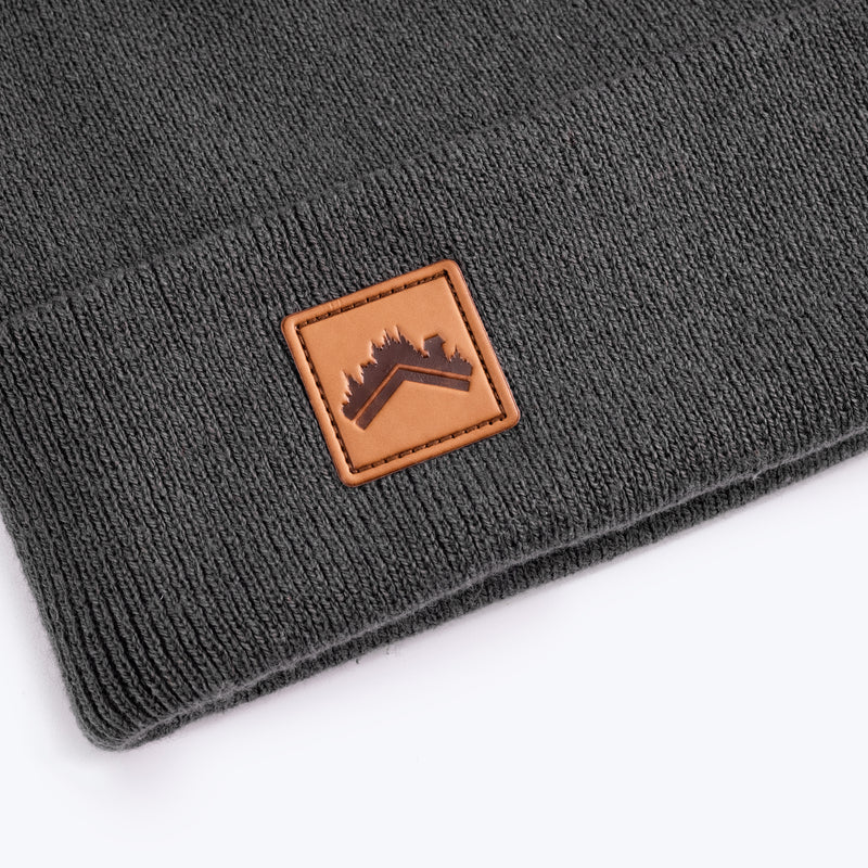 A close up detail shot of a soft dark stone grey 6 panel beanie with a custom ALDRI SUR leather logo patch square stitched to the bottom left fold