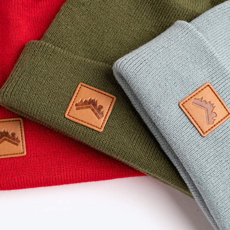 A close up detail trio flat lay shot of the ALDRI SUR recycled acrylic beanie in Spice Red, recycled acrylic beanie in Moss Green, and cotton acrylic blend in soft Glacier Blue. All have custom leather logo patch squares in the bottom left corner of the fold