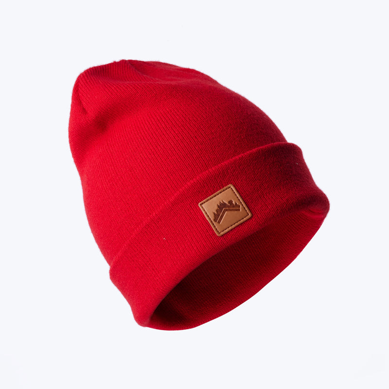 A floating head style shot of a soft spice red recycled acrylic 6 panel beanie with a custom ALDRI SUR leather logo patch square stitched to the bottom left fold